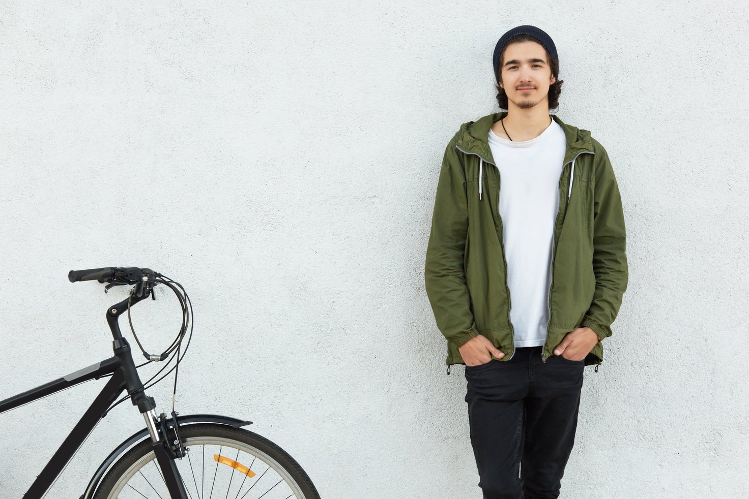 Horizontal view of stylish man keep hands in pockets of black trousers wears green jacket, stands near his bicycle, likes cycling, isolated over white concrete wall. People, youth and teenagers